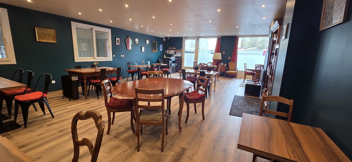 La salle en enfilade vers le comptoir, éclairée par la vitrine sur la rue, avec des tables dressées et des rideaux rouges.