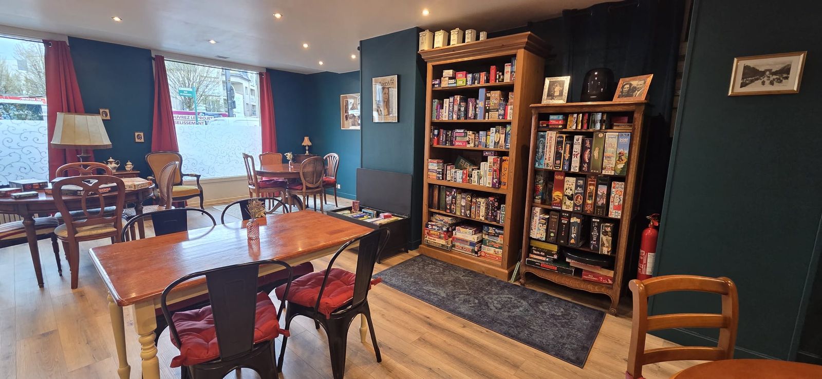 La salle du Manoir Ludique : deux hautes bibliothèques remplies de boîtes de jeux, des tables en bois et des chaises à coussins rouges, sous des murs bleu canard.